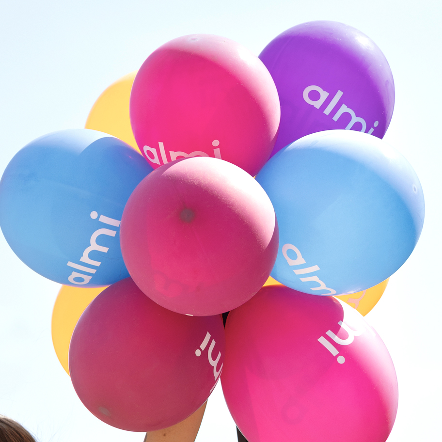 En bunt färgglada ballonger i rosa, blått, lila och gult med texten "almi" tryckt i vitt på ytan. Ballongerna hålls upp mot en klar himmel, vilket skapar en festlig och positiv känsla.