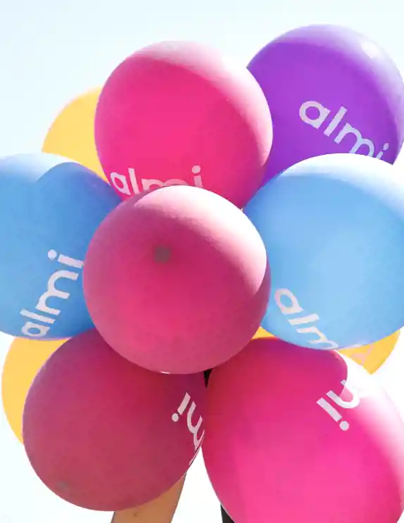 En bunt färgglada ballonger i rosa, blått, lila och gult med texten "almi" tryckt i vitt på ytan. Ballongerna hålls upp mot en klar himmel, vilket skapar en festlig och positiv känsla.
