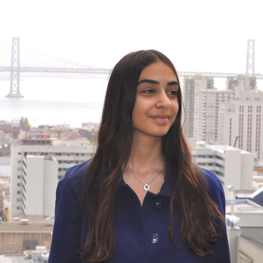 Porträtt av en kvinna med långt mörkt hår, klädd i en mörkblå kappa och ett silverhalsband, stående på en balkong med en stadsvy i bakgrunden. En bro syns i fjärran.