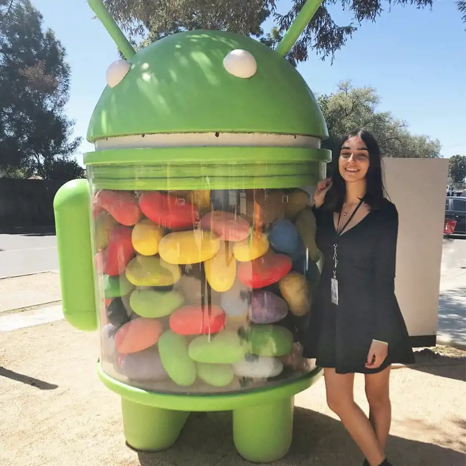 En kvinna i svart klänning med en ID-bricka poserar bredvid en stor Android Jelly Bean-statyn utomhus. Statyn föreställer den gröna Android-maskoten med en genomskinlig mage fylld med färgglada godisbönor. Soligt väder och träd i bakgrunden.