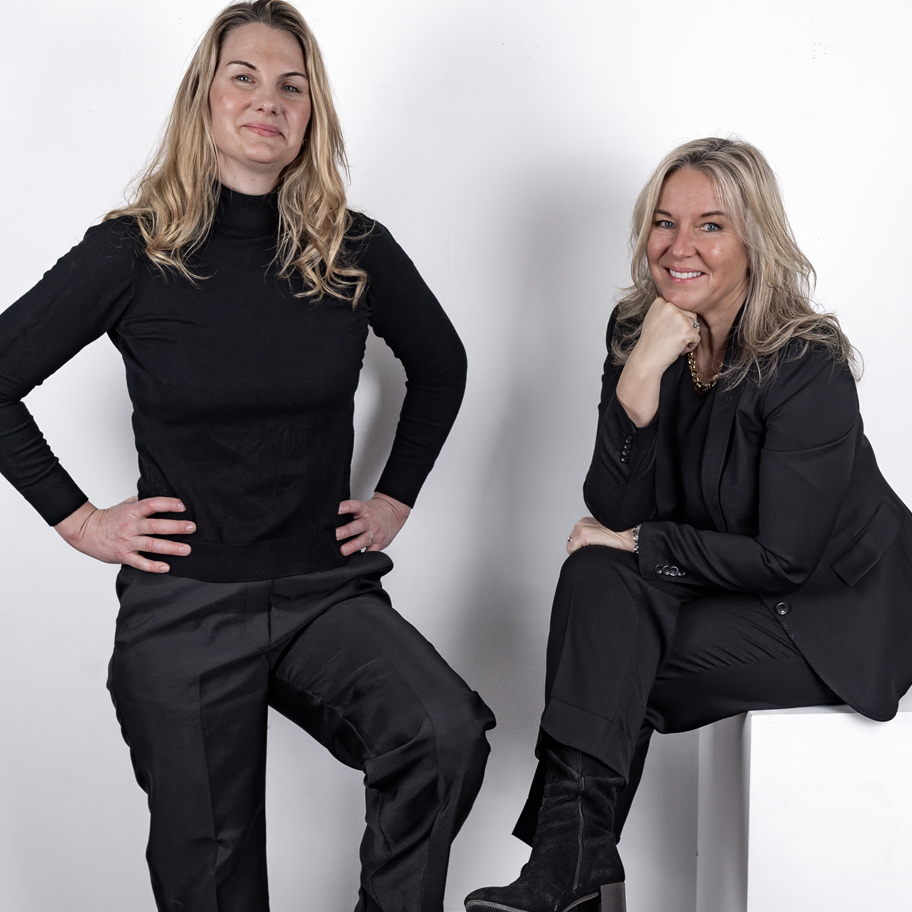 Två kvinnor med blont hår och svarta kläder poserar mot en vit bakgrund. Den ena står med ena handen på höften och ett självsäkert uttryck, medan den andra sitter på en vit kub, lutar handen mot hakan och ler.