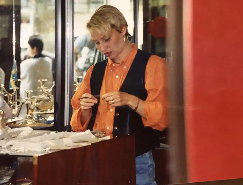 En kvinna med kort blont hår och en orange skjorta med en svart väst står vid en smyckesdisk och inspekterar ett smycke med en pincett. Hon har ett koncentrerat uttryck och flera smycken på händerna. Bakom henne syns en spegelvägg och glasmontrar fyllda med silverföremål, samt en suddig reflektion av människor i bakgrunden.