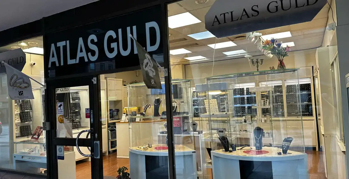 Bild av smyckesbutiken "Atlas Guld" med en svart skylt ovanför entrén. Butiken har stora glasfönster som visar smyckesmontrar fyllda med halsband, ringar och andra smycken. Inuti syns ljusa trädetaljer, speglar och belysning som framhäver smyckena. Utanför hänger en flagga med en bild på ringar.