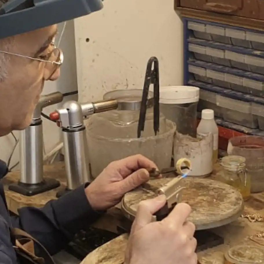 En guldsmed arbetar vid sin arbetsbänk med att värma en ring med en blåslampa. Han bär skyddsglasögon och ett förstoringsglas på huvudet. Arbetsbänken är fylld med verktyg, små burkar och behållare, samt en roterande platta där ringen placeras. Bakom honom finns en hylla med lådor för förvaring av material och verktyg.