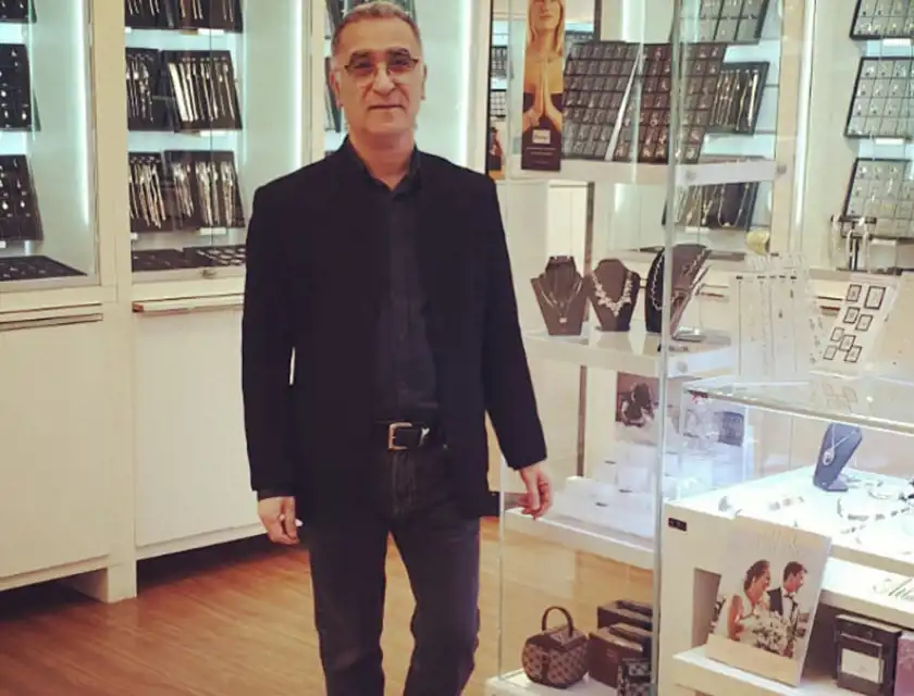 En man i svart kavaj och mörka jeans står i en smyckesbutik. Han har glasögon och ett vänligt uttryck. Bakom honom syns glasmontrar fyllda med halsband, ringar och andra smycken, samt en affisch med en kvinna som håller händerna i en böneliknande gest.