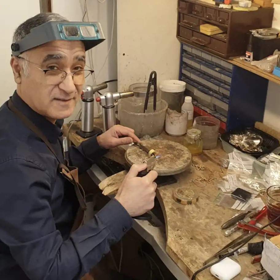 En guldsmed sitter vid sin arbetsbänk och arbetar med att värma en ring med en blåslampa. Han bär en mörkblå skjorta, ett förstoringsglas på huvudet och ett brunt förkläde. Han tittar mot kameran med ett vänligt uttryck. Arbetsbänken är fylld med verktyg, guldsmycken, små burkar och plastpåsar med märkningar. I bakgrunden finns en hylla med lådor för förvaring av material och verktyg.