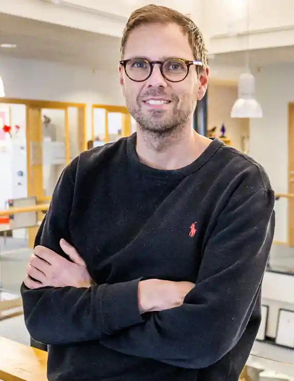 En man med kort brunt hår, skäggstubb och glasögon ler mot kameran. Han bär en svart tröja med en röd Polo Ralph Lauren-logga och har armarna i kors. Bakgrunden visar en modern kontorsmiljö med glasräcken, trädetaljer och hängande lampor. Dörrar och glasfönster i bakgrunden leder till olika rum i byggnaden.