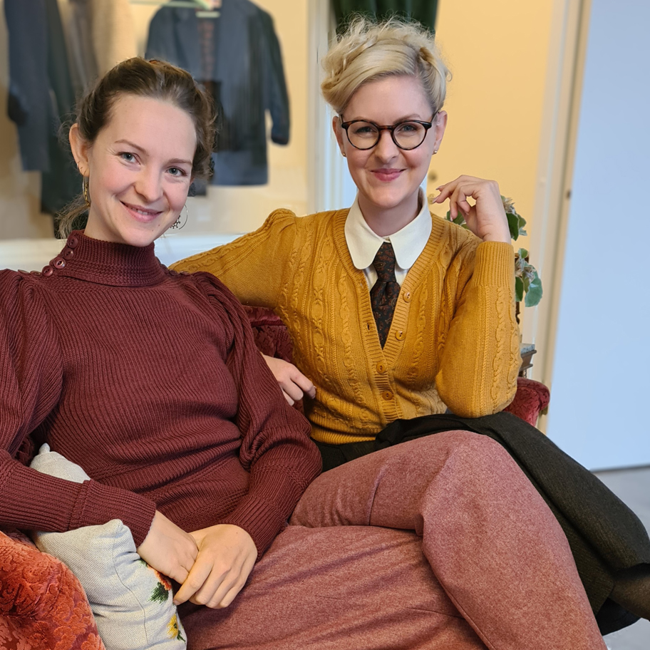 Två kvinnor sitter avslappnat i en röd sammetsfåtölj och ler mot kameran. Den ena bär en vinröd ribbstickad tröja med puffärmar, medan den andra har en senapsgul kabelstickad kofta över en vit kragskjorta med slips. Hon har blont, uppsatt hår och glasögon. Bakgrunden visar en interiör med kläder upphängda på väggen och en dörröppning till ett annat rum.