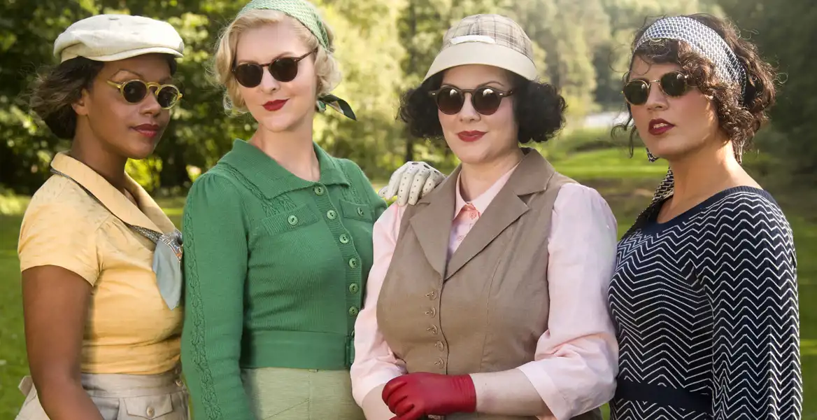 Fyra kvinnor står utomhus i en grönskande park, klädda i eleganta retrokläder inspirerade av 1940- och 1950-talet. De bär solglasögon, hattar, hårband och handskar, och har djärva röda läppstift. En kvinna i gul blus och grå kjol har en snusnäsduk knuten vid halsen. En annan i grön kofta och matchande hårband ler subtilt. Den tredje bär en beige väst över en rosa blus, med röda handskar och en rutig keps. Den fjärde kvinnan har mörkt lockigt hår och bär en marinblå klänning med sicksackmönster och ett mönstrat hårband. De poserar självsäkert och elegant.