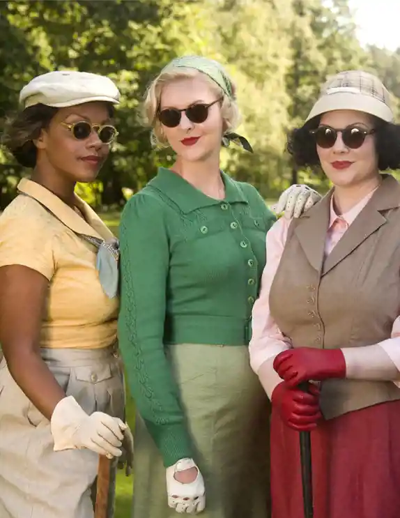Fyra kvinnor står utomhus i en grönskande park, klädda i eleganta retrokläder inspirerade av 1940- och 1950-talet. De bär solglasögon, hattar, hårband och handskar, och har djärva röda läppstift. En kvinna i gul blus och grå kjol har en snusnäsduk knuten vid halsen. En annan i grön kofta och matchande hårband ler subtilt. Den tredje bär en beige väst över en rosa blus, med röda handskar och en rutig keps. Den fjärde kvinnan har mörkt lockigt hår och bär en marinblå klänning med sicksackmönster och ett mönstrat hårband. De poserar självsäkert och elegant.