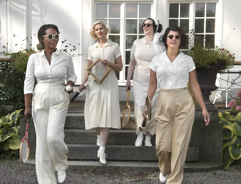 Fyra kvinnor klädda i eleganta, vintageinspirerade vita och beige sportkläder från tidigt 1900-tal går nerför trappan till en herrgårdsliknande byggnad med stora fönster. De bär solglasögon och håller i gamla tennisracketar och en baseboll. Alla har en självsäker och stilfull hållning. Bakgrunden är en vacker trädgård med gröna växter och blomkrukor.