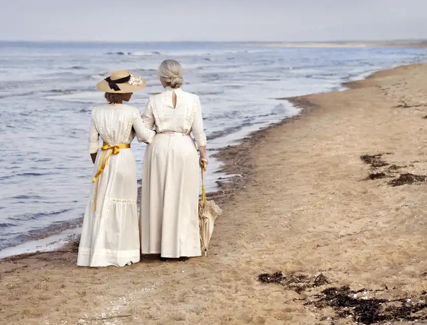 Två kvinnor klädda i ljusa, vintageinspirerade klänningar från tidigt 1900-tal promenerar längs en sandstrand med havet i bakgrunden. Den ena kvinnan bär en bredbrättad hatt med ett svart band och en gul sidenrosett runt midjan, medan den andra har uppsatt grått hår och håller ett parasoll i handen. De går arm i arm och blickar ut över havet, vilket skapar en känsla av nostalgi och lugn.
