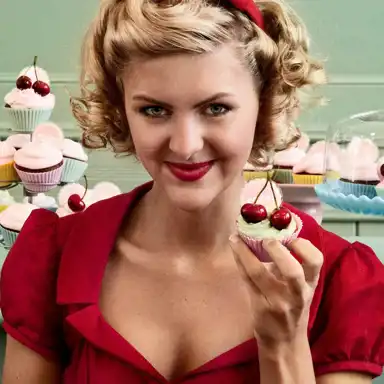 En kvinna med blont lockigt hår, ett rött hårband och en röd klänning ler lekfullt medan hon håller upp en cupcake dekorerad med ljusgrön frosting och två körsbär. Hennes makeup inkluderar röda läppar och en subtil vintage-look. I bakgrunden syns ett bord fyllt med färgglada cupcakes, många dekorerade med rosa frosting och körsbär, uppställda på ett kakställ. Väggarna är målade i en pastellgrön nyans, vilket skapar en retroinspirerad och inbjudande atmosfär.