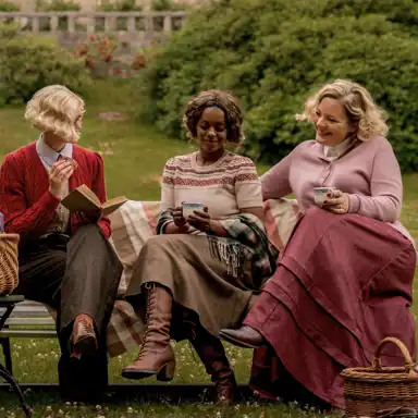 Tre kvinnor i vintageinspirerade höstkläder sitter på en parkbänk utomhus, insvepta i en filt, och dricker te ur mönstrade koppar. Den ena bär en röd kofta över en skjorta och slips, håller en bok och en kaka. Kvinnan i mitten har en stickad tröja med nordiskt mönster, en yllekjol och höga snörstövlar, och är insvept i en rutig sjal. Den tredje kvinnan bär en rosa stickad tröja och en lång röd kjol, och ler medan hon pratar med de andra. Bredvid bänken står en korg, och i bakgrunden syns grönska och en stenmur, vilket skapar en mysig och nostalgisk atmosfär.