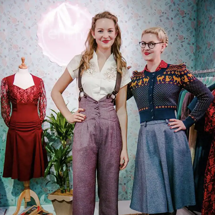 Två kvinnor med vintageinspirerad stil står framför en retroinredd bakgrund med blommigt tapetmönster. Den ena bär en vit blus med broderier, höga lila byxor med hängslen och har håret uppsatt i mjuka lockar. Den andra har en blå kjol och en mönstrad kofta med röd skjorta under, samt glasögon och en flätad frisyr. De poserar med händerna på höfterna och ler. Bakom dem syns en provdocka med en röd spetsklänning, samt klädställningar med fler retrokläder.