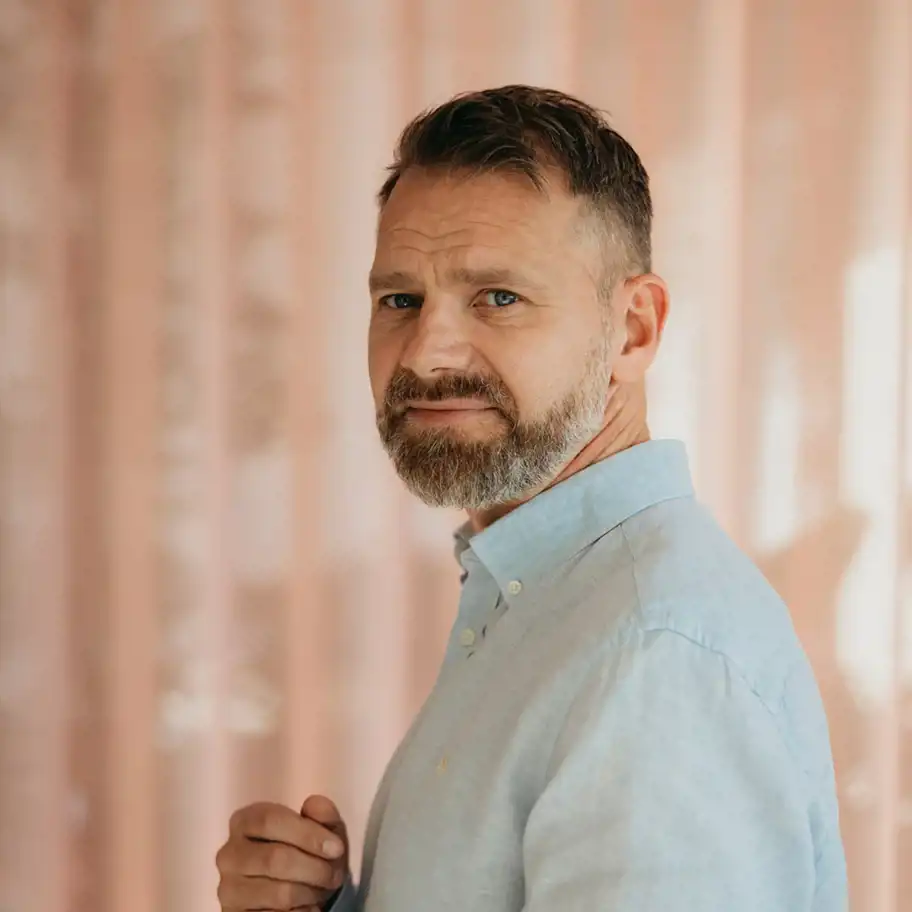 En medelålders man med kortklippt hår och vältrimmat skägg tittar in i kameran med ett lugnt och eftertänksamt uttryck. Han bär en ljusblå skjorta och står framför en bakgrund med mjuka rosa gardiner, vilket skapar en varm och harmonisk känsla i bilden. Ljuset är mjukt och naturligt, vilket framhäver ansiktets detaljer.