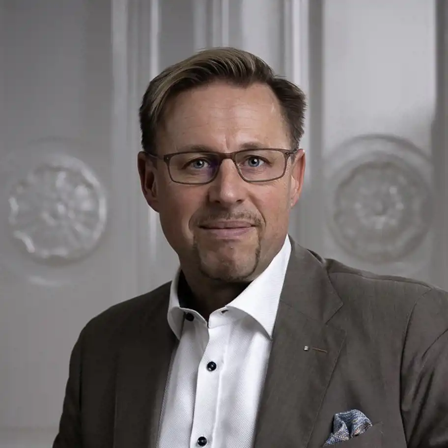 En man med kort hår och glasögon ler lätt mot kameran. Han bär en brun kostym med en vit skjorta och en blå mönstrad näsduk i bröstfickan. Bakgrunden består av en elegant, vit panelvägg med dekorativa detaljer, vilket ger en stilren och professionell känsla.