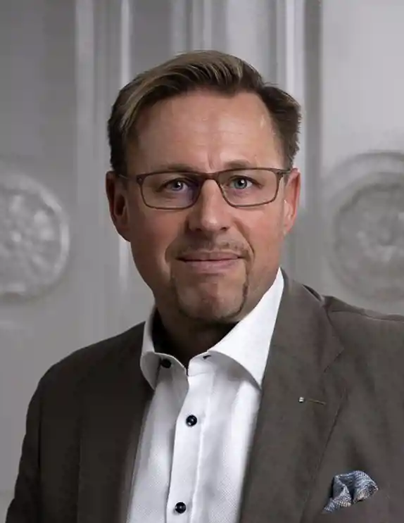 En man med kort hår och glasögon ler lätt mot kameran. Han bär en brun kostym med en vit skjorta och en blå mönstrad näsduk i bröstfickan. Bakgrunden består av en elegant, vit panelvägg med dekorativa detaljer, vilket ger en stilren och professionell känsla.