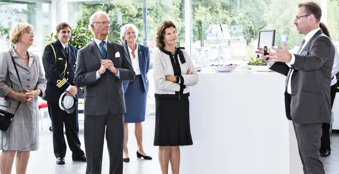 En man i kostym håller en presentation för en grupp personer i ett ljust, modernt rum med stora fönster. Bland åhörarna finns kungen i mörk kostym och glasögon samt drottning Silvia i en ljus jacka och svart klänning, som båda lyssnar uppmärksamt. I bakgrunden syns ytterligare personer, varav en i en marinblå uniform med gulddetaljer håller en vit mössa. På ett vitt bord framför presentatören står skålar med sallad och bär samt en uppställd skärm. Utanför fönstren syns grönska och en parkeringsplats.