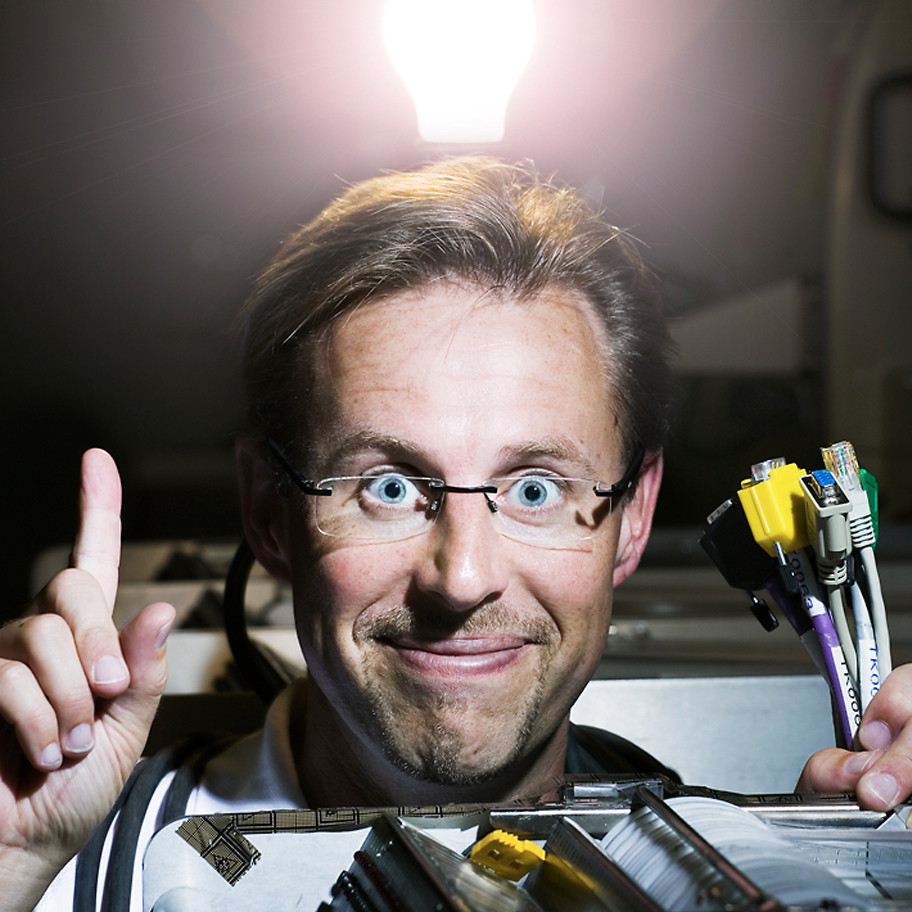 En man med glasögon och uppspärrade ögon håller upp en bunt nätverkskablar i ena handen och pekar uppåt med den andra. Bakom honom lyser en glödlampa starkt, vilket skapar en dramatisk belysning och får det att se ut som att han fått en idé. Han bär en ljus skjorta och är omgiven av teknisk utrustning, inklusive kretskort och kablar. Bakgrunden är något mörk och suddig, vilket lägger fokus på mannens ansikte och uttryck.