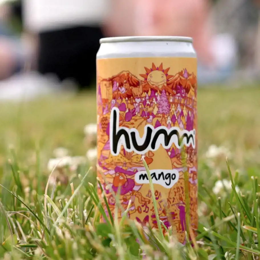 En färgglad burk med designen "humm mango" står i gräset bland små vita blommor. I bakgrunden syns suddiga människor som sitter på en filt, vilket skapar en avslappnad picknickkänsla.
