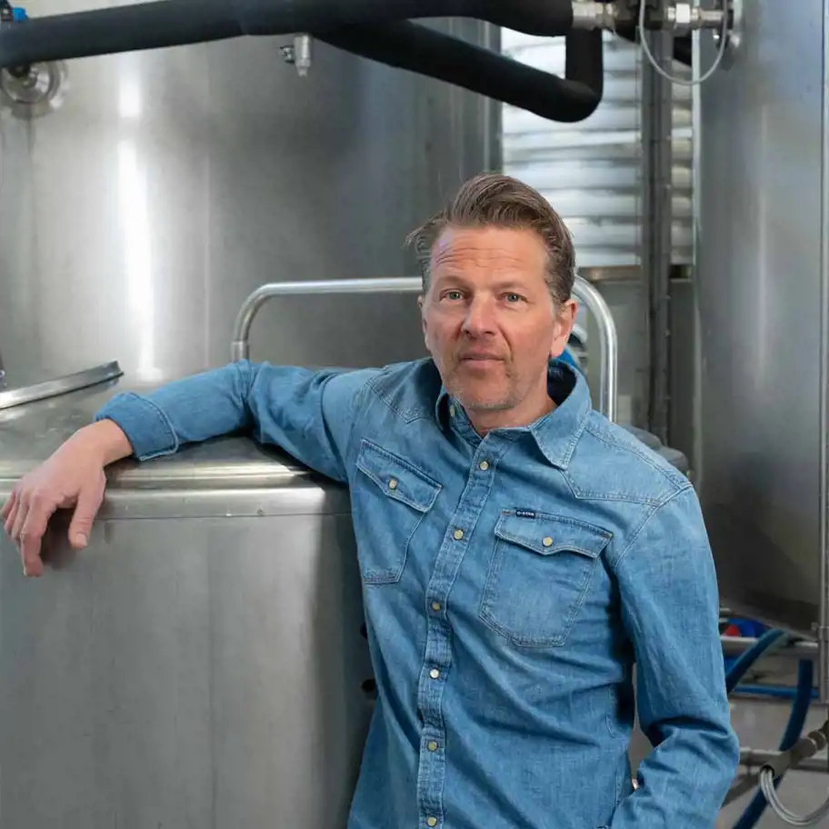 En man med kort skägg och bakåtkammat hår står lutad mot en stor rostfri tank i en industriell miljö. Han bär en blå jeansskjorta. Bakgrunden består av fler metalltankar, rör och annan utrustning, vilket antyder att han befinner sig i en produktionsanläggning, möjligen ett bryggeri eller en livsmedelsfabrik.