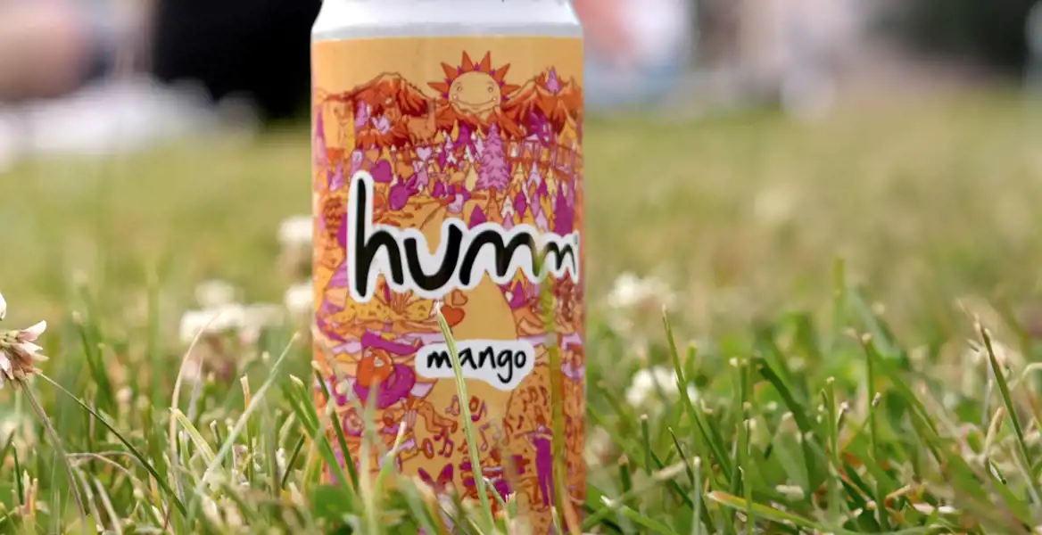 En färgglad burk med designen "humm mango" står i gräset bland små vita blommor. I bakgrunden syns suddiga människor som sitter på en filt, vilket skapar en avslappnad picknickkänsla.