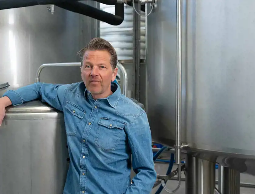 En man med kort skägg och bakåtkammat hår står lutad mot en stor rostfri tank i en industriell miljö. Han bär en blå jeansskjorta. Bakgrunden består av fler metalltankar, rör och annan utrustning, vilket antyder att han befinner sig i en produktionsanläggning, möjligen ett bryggeri eller en livsmedelsfabrik.
