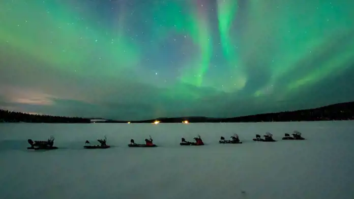 En grupp snöskotrar står parkerade på en vidsträckt frusen sjö under en mörk natthimmel. Ovanför dem dansar ett spektakulärt norrsken i gröna och blå nyanser, vilket skapar en magisk och drömsk atmosfär. I horisonten syns en tät skog och några avlägsna ljuspunkter, som antyder närvaron av en bosättning eller läger.