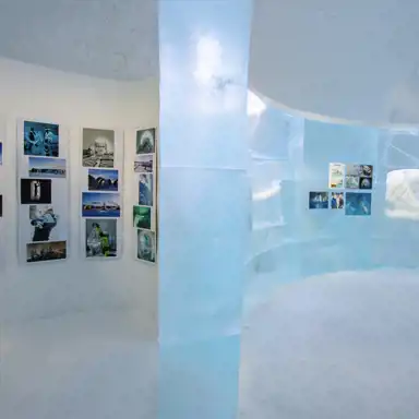 Ett konstgalleri inuti en isbyggnad, där genomskinliga blå isväggar elegant separerar rummet. Fotografier och informationsskyltar är uppsatta på de vita väggarna, vilket ger en inblick i ishotellets historia och design. Det reflekterande ljuset från isen skapar en magisk och kall atmosfär, medan den böjda strukturen på väggarna förstärker den unika arkitekturen i denna frusna utställning.