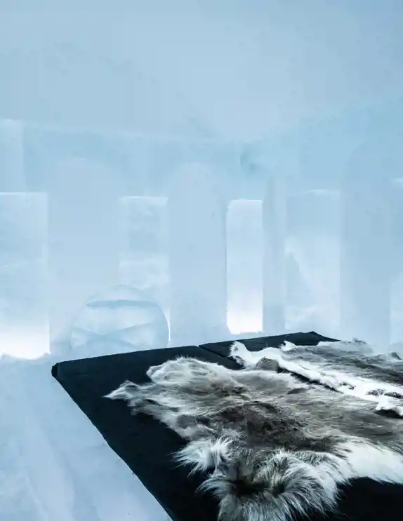 Ett rum i ett ishotell där väggar, tak och möbler är helt gjorda av is och snö. Sängen, placerad på en isig plattform, är täckt med tjocka renfällar för att ge värme och komfort. Väggarna är dekorerade med vackert utformade valvbågar, och mjuk belysning skapar en magisk och lugnande atmosfär. En stor isskulptur i form av en klotliknande struktur står vid sidan av rummet, vilket förstärker den unika och konstnärliga känslan.