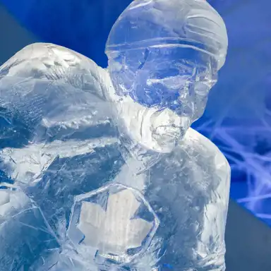 En detaljerad isskulptur av ishockeyspelaren Börje Salming, med hjälm och en intensiv, fokuserad min. Spelarens tröja har ett ingraverat lönnlövs-emblem, vilket antyder en koppling till Kanada. Skulpturen är skickligt uthuggen med skarpa linjer och texturer som förstärker intrycket av rörelse och styrka. Den omgivande blå belysningen skapar en frostig, nästan magisk atmosfär, där isens genomskinlighet framhävs och detaljerna får liv.