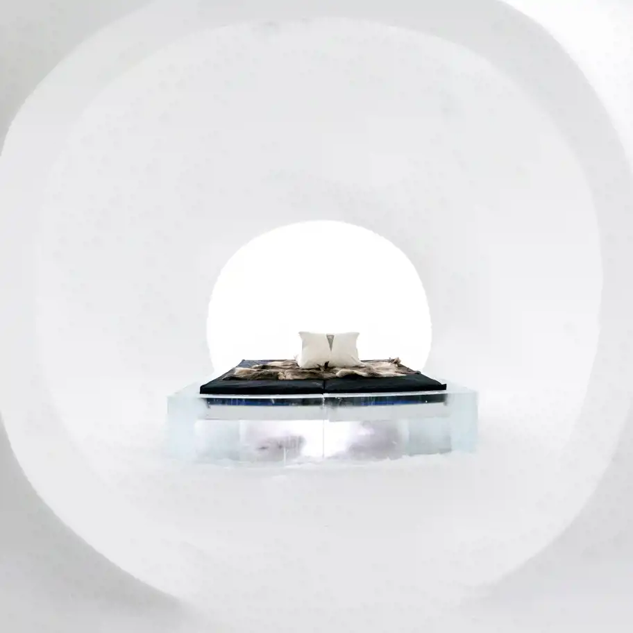 En minimalistisk sovplats i ett isskulpterat rum med mjukt rundade väggar och tak. I mitten står en säng på en transparent isplattform, täckt med mörka filtar och fällar samt en enkel vit kudde. Rummets ljusa och rena estetik förstärks av den starka kontrasten mellan den svarta sängklädseln och det nästan lysande vita rummet. Öppningen i bakgrunden ger en känsla av rymd och lugn, som om sängen svävar i en stilla, frusen värld.