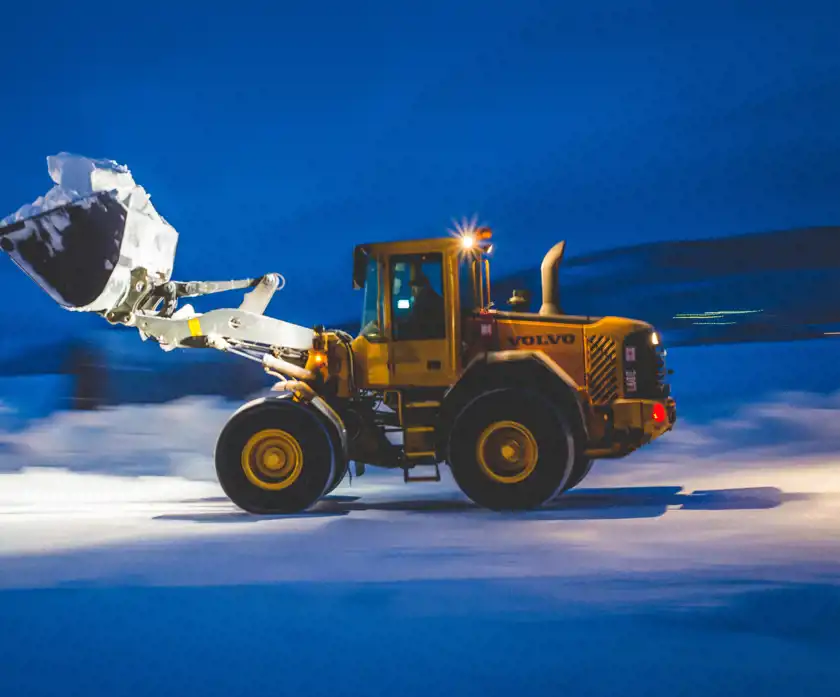 En gul Volvo hjullastare i rörelse genom ett snölandskap under den mörkblå natthimlen. Skopan är fylld med snö och lyfts uppåt medan fordonets kraftfulla däck greppar den frusna marken. Föraren syns genom hyttens fönster, fokuserad på sitt arbete. Bakgrundens suddiga linjer ger en känsla av hastighet och effektivitet i den kalla, nordliga miljön.