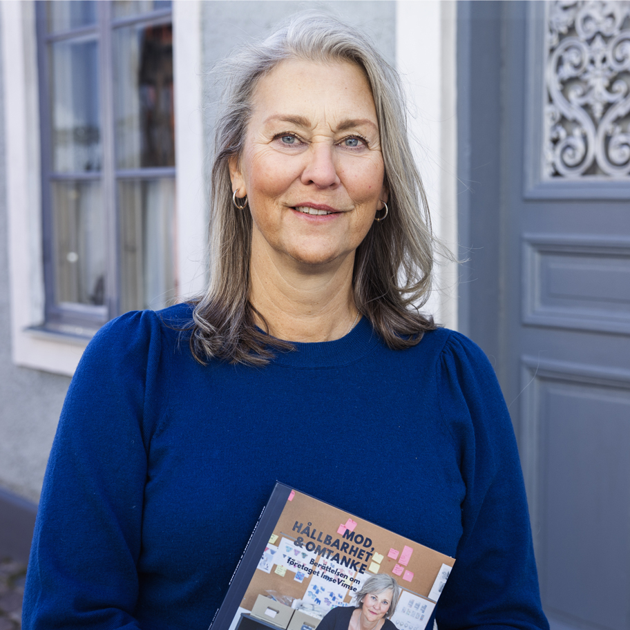 En kvinna med axellångt hår står utomhus framför en grå dörr och ett fönster. Hon har på sig en mörkblå tröja och håller en bok i handen med titeln "MOD, HÅLLBARHET & OMTANKE". Kvinnan ler vänligt och tittar in i kameran. Bakgrunden består av en ljus fasad med ett dekorerat dörrparti.
