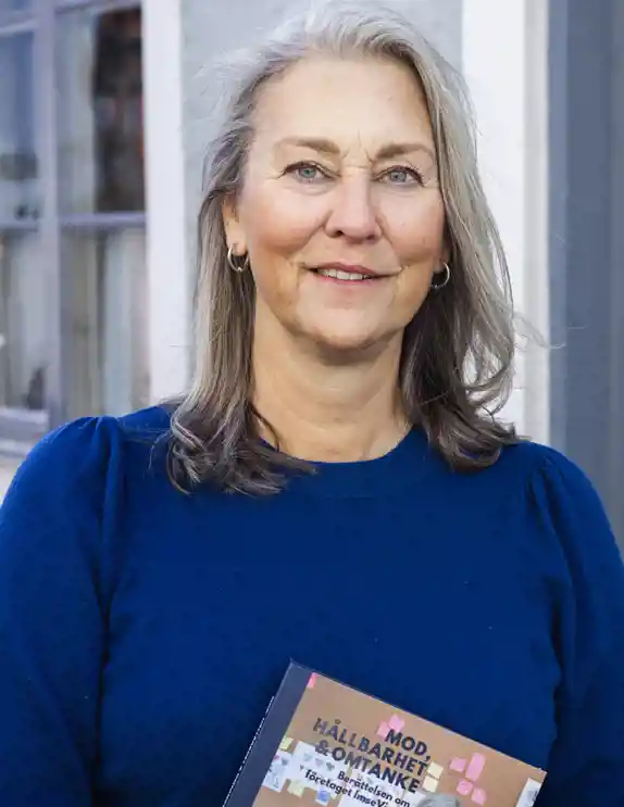En kvinna med axellångt hår står utomhus framför en grå dörr och ett fönster. Hon har på sig en mörkblå tröja och håller en bok i handen med titeln "MOD, HÅLLBARHET & OMTANKE". Kvinnan ler vänligt och tittar in i kameran. Bakgrunden består av en ljus fasad med ett dekorerat dörrparti.