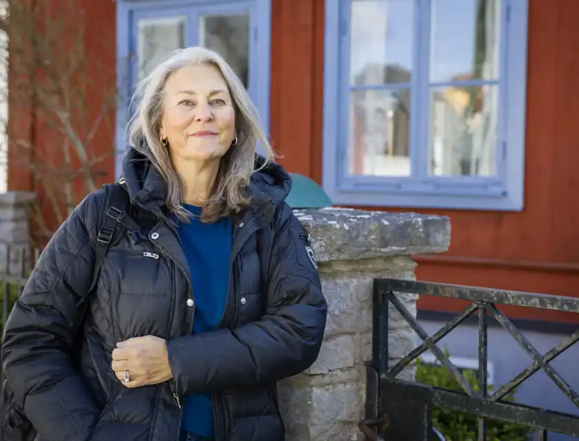 En kvinna i en svart vadderad jacka och en blå tröja står utomhus vid en stenmur och en svart järngrind. Hon har en ryggsäck över ena axeln och håller ena handen på jackan. Bakom henne syns en röd träbyggnad med blå fönsterkarmar och en speglande ruta. Det är en solig dag, och några grenar syns i bakgrunden.