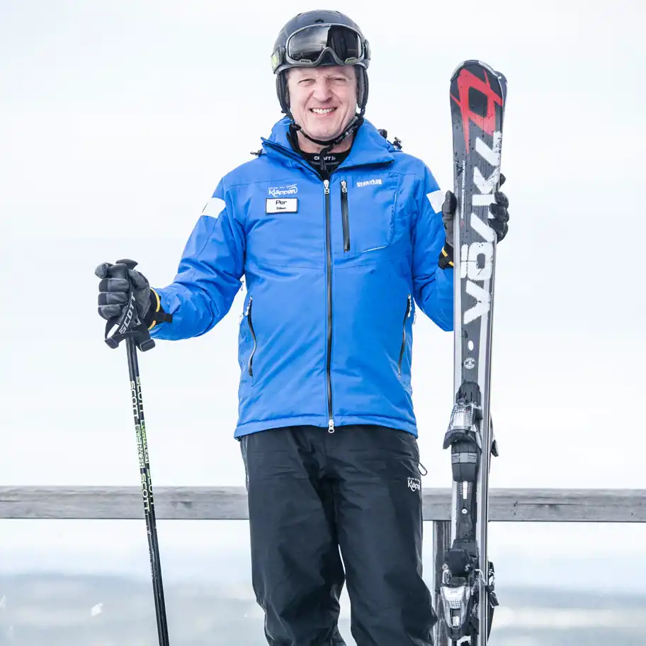 En man i en blå skidjacka med namnskylten "Per, Sälen" står utomhus i en vintermiljö. Han håller en skidstav i ena handen och en svart och röd slalomskida i den andra. Han bär svarta skidbyxor, handskar, en svart hjälm och skidglasögon. Bakgrunden är ljus och snötäckt.