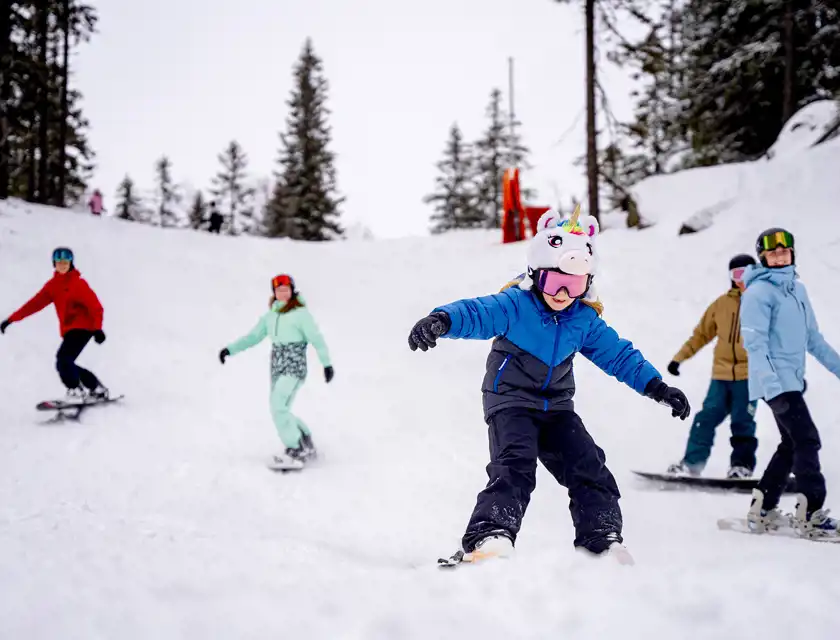 En grupp snowboardåkare åker nerför en snötäckt backe omgivna av träd. I förgrunden syns ett barn i en blå jacka, svarta skidbyxor och en hjälm med en enhörningsmössa. Bakom barnet åker flera personer i färgglada skidkläder, inklusive en person i en röd jacka och en annan i en mintgrön overall. Himlen är molnig och snön täcker både backen och omgivande berg.