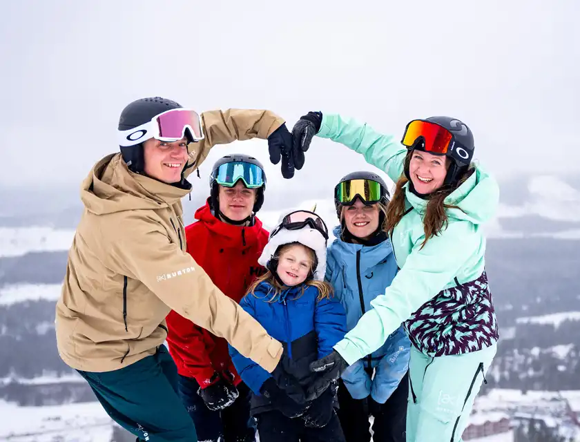 En grupp på fem personer står på en snötäckt bergstopp och formar ett hjärta med sina händer. De är klädda i färgglada skid- och snowboardkläder, inklusive en person i en beige jacka, en i en röd jacka och en i en mintgrön overall. I mitten står ett barn i en blå jacka och en mössa med enhörningsdetaljer. Alla ler och ser glada ut. Bakgrunden visar ett vintrigt bergslandskap med snöklädda sluttningar och dalar.
