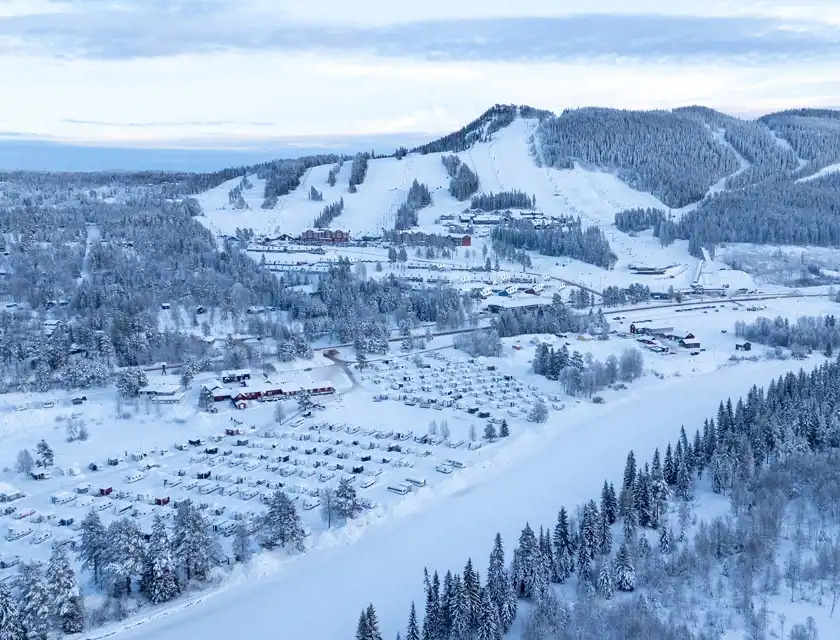 Ett vinterlandskap sett från ovan med snötäckta skidbackar, skog och en dal med byggnader och ett campingområde fullt av husvagnar. Backarna är omgivna av skog och små stugor, och skidspår slingrar sig nerför bergen. Himlen har en mjuk blå ton, och snön reflekterar det kalla ljuset.