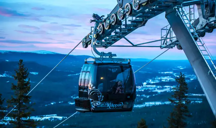 En gondollift från Kläppen svävar högt ovanför ett vinterlandskap i skymningsljus. Himlen har blå och rosa toner, och snötäckta berg och skogar breder ut sig i bakgrunden. Gondolen är svart med vita dekorationer, och en person i färgglada vinterkläder syns genom fönstret. Lyftstationens metallkonstruktion och vajrar är tydligt synliga i bilden.