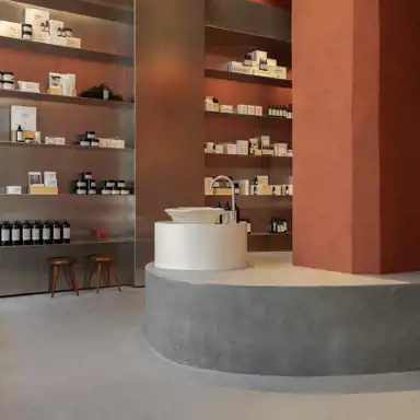 En stilren butik med hyllor fyllda med hudvårdsprodukter i minimalistiska förpackningar. Inredningen har en varm färgpalett med rostfärgade väggar, rostfria hyllor och en grå betongplattform. På plattformen står ett runt handfat i sten med en modern kran. Två träpallar och en liten röd dekorhäst syns vid en av hyllorna.