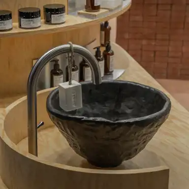 Ett stilrent handfat i mörk keramik med en ojämn yta, placerat på en ljus träbänk. En böjd metallkran är monterad bredvid, med en vit tvål upphängd i ett snöre. I bakgrunden syns hyllor med hudvårdsprodukter i bruna glasflaskor och svarta burkar, samt en vägg i rödbrunt tegel.