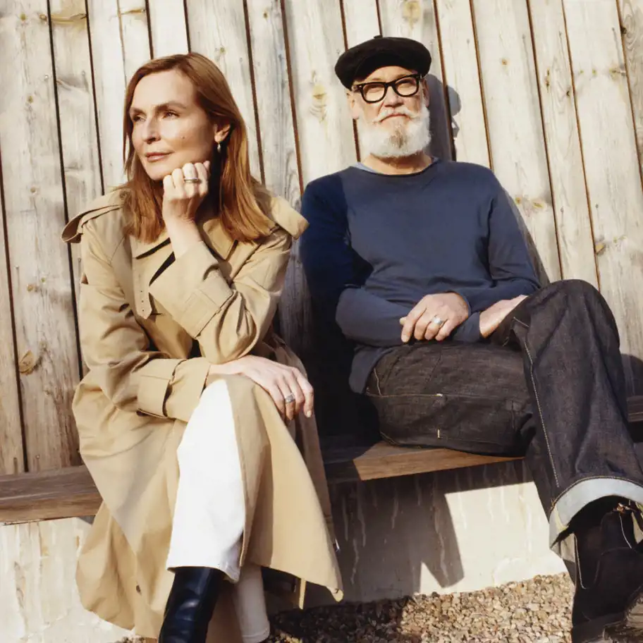 Monica Kylén och Mats Johansson, grundare av L:a Bruket. En kvinna och en man sitter på en bänk mot en vägg av träplankor i solljus. Kvinnan bär en beige trenchcoat, vita byxor och svarta stövlar, och lutar sig lätt fram med ena handen vid ansiktet. Mannen bär en mörkblå långärmad tröja, mörka jeans, en svart keps och glasögon, och har armarna avslappnat korsade. De ser ut att vara i en lugn och avslappnad stund utomhus.