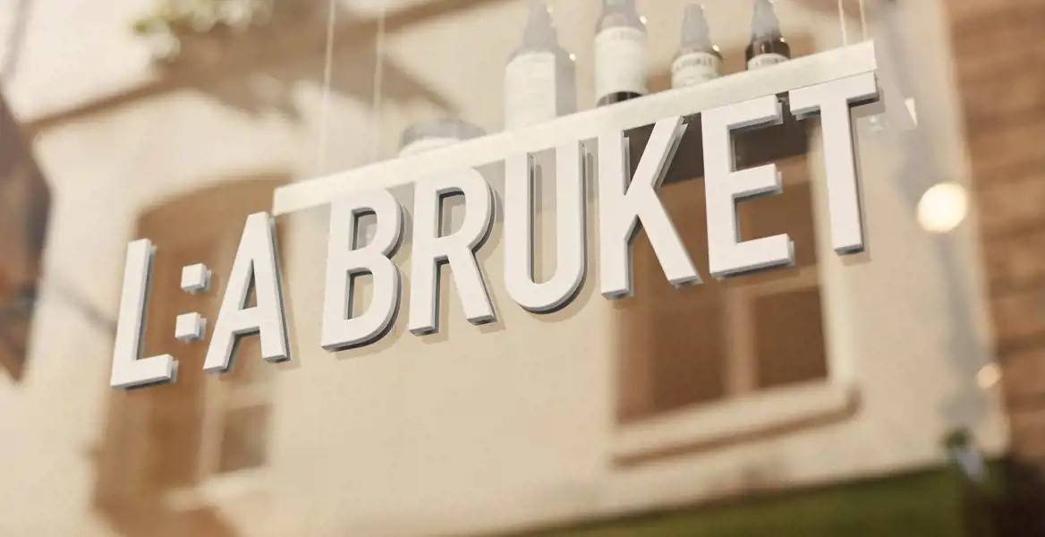 Skyltfönster till L:a Bruket i Paris, Marais. Närbild av en skylt med texten "L:A BRUKET", monterad på ett glasfönster. Skylten är i vita, upphöjda bokstäver. I bakgrunden syns reflektioner av en byggnad med fönster och ljusa fasader, samt några hudvårdsprodukter på en hylla innanför glaset. Ljuset är varmt och naturligt.