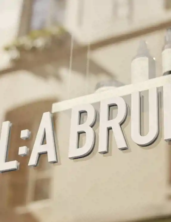 Skyltfönster till L:a Bruket i Paris, Marais. Närbild av en skylt med texten "L:A BRUKET", monterad på ett glasfönster. Skylten är i vita, upphöjda bokstäver. I bakgrunden syns reflektioner av en byggnad med fönster och ljusa fasader, samt några hudvårdsprodukter på en hylla innanför glaset. Ljuset är varmt och naturligt.