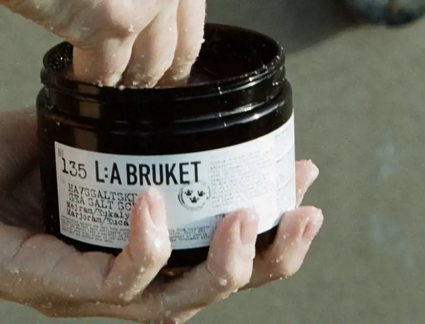 En närbild av en hand som tar produkt ur en svart burk med etiketten "L:A BRUKET No 135 HAVSSALTSKRUBB". Händerna är fuktiga och täckta av små saltkorn. Bakgrunden är suddig och ser ut att vara en naturlig utomhusmiljö.