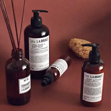 En samling bruntonade flaskor från L:A BRUKET med hud- och kroppsvårdsprodukter står på en rostfärgad yta. Produkterna inkluderar tvål, body lotion och doftspridare med etiketter i svart och vitt. En naturlig tvättsvamp ligger bredvid flaskorna, och en av dem har doftpinnar i sig. Stilen är minimalistisk och harmonisk.