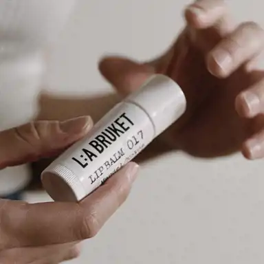 En närbild av en person som håller i ett vitt läppbalsam från L:A BRUKET med texten "LIP BALM 017" på etiketten. Händerna är mjukt belysta, och bakgrunden är suddig med en neutral färgton. Stilen är minimalistisk och naturlig.